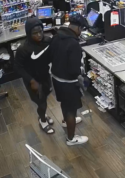Camera photo of 2 suspects inside store. First suspect is wearing a black hoodie with a large white Nike symbol on front, black shorts, white socks and black slip on sandals. Second suspect is wearing black hoodie and shorts, white Nike shoes with black check mark and a newsboy hat. 